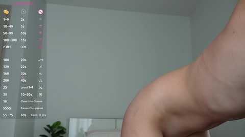 Video of a nude, light-skinned person in a bedroom, with a smartphone displaying a fitness app screen on the left. The background shows a simple, white-walled room with minimal decor.