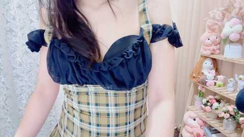 Video of an Asian woman with long black hair wearing a plaid dress with a ruffled neckline, standing in a room filled with plush toys and a wooden ladder shelf.