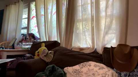 Media: A video of a cozy bedroom with a bed covered in a dark brown blanket, a yellow stuffed toy, and a beige hat. Soft light filters through sheer curtains, casting a warm glow.
