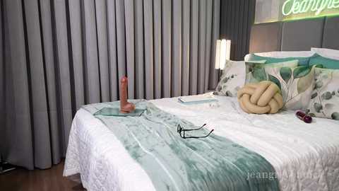 Video of a modern, minimalist bedroom with gray curtains, a white bed, teal blanket, and a realistic penis-shaped candle on a nightstand.