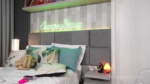 Video of a modern bedroom with a grey upholstered headboard, white bedding, teal and beige pillows, a neon \"Jenney Sman\" sign, and a cozy nightstand with a candle and a pink book.