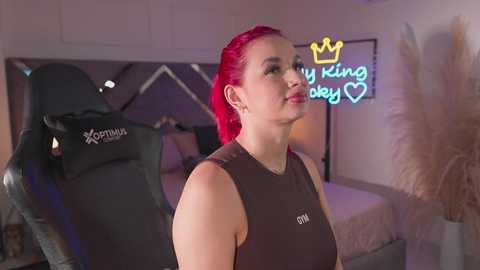 Video of a fair-skinned woman with red hair, wearing a black sports bra, standing in a modern bedroom with a black gaming chair and a neon sign reading \"my king, baby, I love you.\