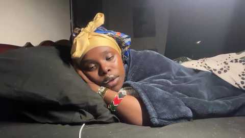 Video of a sleeping Black woman with dark skin, wearing a yellow headwrap, blue blanket, and red bracelet. She lies on a bed with black and white sheets in a dimly lit room.