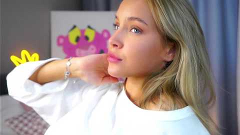 Video of a young Caucasian woman with light skin and straight blonde hair, wearing a white top. She has a relaxed expression, resting her chin on her hand. Background features a colorful, cartoonish painting of a frog.