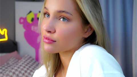 Video of a young Caucasian woman with fair skin, long blonde hair, and blue eyes, wearing a white robe, looking contemplative. Background features a bed with a pink bear and a yellow crown.