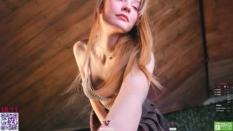 A video of a young, fair-skinned woman with long, straight blonde hair, wearing a beige knitted top and plaid skirt, leaning forward, eyes closed, in a rustic wooden room.