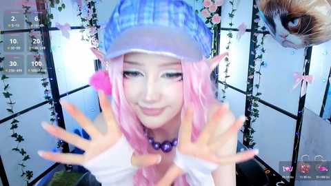 Video of a young Asian woman with pink hair, wearing a blue baseball cap, pointy elf ears, and a purple necklace, smiling, with virtual camera effects, in a room with hanging flowers and a cat in the background.