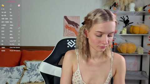 Video of a young Caucasian woman with light blonde hair and fair skin, wearing a floral-patterned camisole, sitting on a black and white gaming chair in a cozy, autumn-decorated room with pumpkins and a framed deer print.