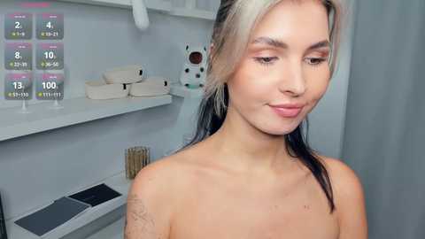 Video of a topless woman with light skin, blonde ombre hair, and a small tattoo on her left arm, standing in a sterile medical room with white walls and medical equipment, including a stethoscope.