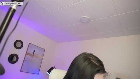 Video of a woman with long black hair sitting in a hair salon, looking away, under fluorescent lighting. White ceiling with circular light fixture. Minimalist decor, including abstract art on the wall.