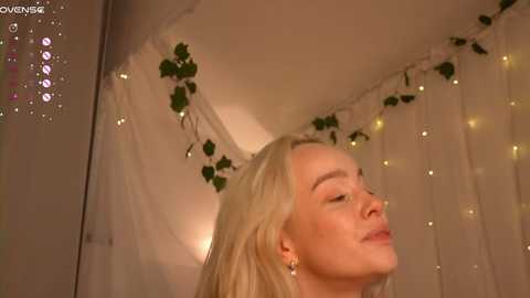 Video of a serene blonde woman with closed eyes, adorned with delicate, dangling earrings, against a softly lit, fairy-lit background.