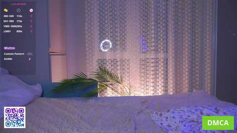 Video of a cozy bedroom with white curtains, a green potted plant, and a blue and white striped pillow on a plush, white bed. A live stream screen on the left shows a camera icon, settings, and a watermark.