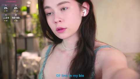 A video of a young woman with long brown hair, wearing a blue lace top, holding a phone, with a blurred background.
