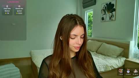 Video of a young woman with long brown hair, wearing a black sheer top, sitting in a minimalist bedroom with white walls, a bed, and a window with greenery.