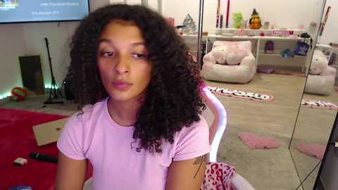 Video of a young Black girl with curly hair, wearing a pink T-shirt, sitting in a cozy living room with a TV, plush toys, and a red couch.