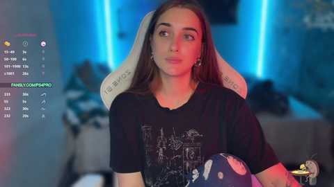 Media: Video of a young woman with long brown hair in a black graphic tee, sitting on a gaming chair, in a dimly lit room with blue lighting.