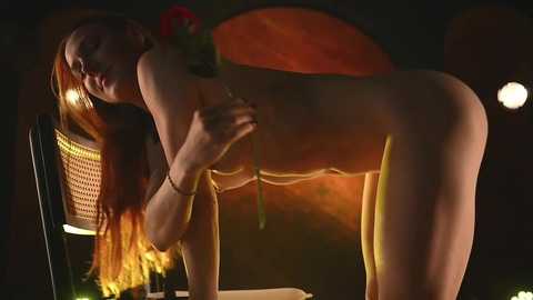 Video of a nude, slender, fair-skinned woman with long red hair, bending over a table, holding a red rose. She wears a gold necklace. Background is dimly lit, with a wicker chair, and warm, orange-toned lighting.
