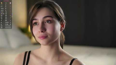 Video of a young, fair-skinned woman with short brown hair, wearing a black spaghetti strap top. Background is blurred, featuring a warm-toned room with a lamp.