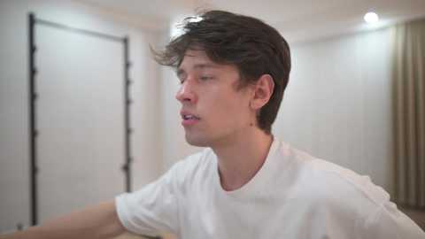 Video of a young Caucasian man with tousled dark hair, wearing a white T-shirt, looking slightly disoriented in a dimly lit room with white walls and beige curtains.