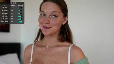 Video of a fair-skinned woman with long brown hair, wearing a light green off-shoulder top, smiling, in a modern bedroom with a white wall and a bed visible in the background.
