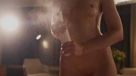 Video of a nude, fair-skinned woman in a dimly-lit room, holding a glass of wine, steam rising from the liquid, blurred background with a chair and plant.