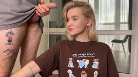 Video of a young woman with light skin and straight blonde hair wearing a \"Not Safe for School\" shirt, kneeling in front of a man lifting his grey shirt to expose his erect circumcised penis. Background shows a modern room with large windows and a chair.