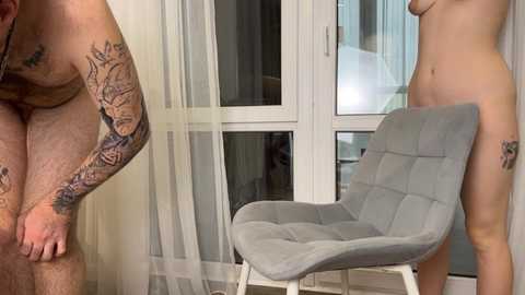 Video of a naked man with tattoos on his arm and chest, standing next to a nude woman with tattoos on her thighs, in a modern, light-filled room with a gray chair and white-framed glass doors.