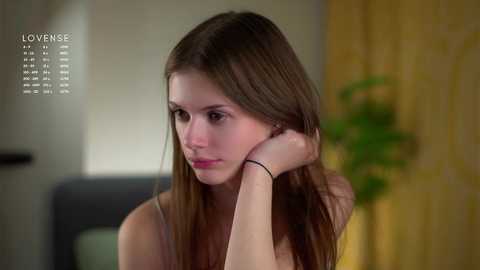 Video of a young woman with long brown hair, light skin, and slender physique, wearing a yellow top, sitting with her head resting on her hand, looking pensive. Background features blurred indoor setting with a potted plant and yellow curtains. Text \"LOVENSE\" appears on the left.