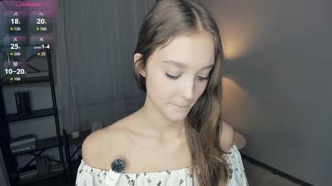 Video of a young Caucasian woman with long brown hair, wearing a white off-shoulder top, looking down with a soft smile. Background includes a digital clock showing 18:20, a bookshelf, and a dimly lit room.