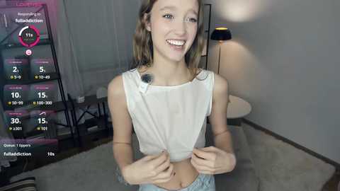 Video of a smiling young white woman with light brown hair, wearing a white crop top and blue pajama pants, standing in a dimly lit bedroom.
