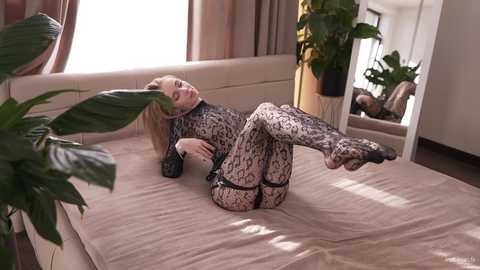 Video of a blonde woman in leopard-print bodysuit lying on beige bed, surrounded by potted plants, mirror, and curtains in a bright, modern room.