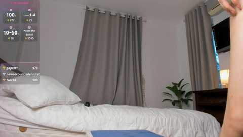 Video of a bedroom with a white bed, gray curtains, and a potted plant. A person, partially visible, is standing near the bed. The image includes a virtual reality overlay.