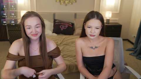 Video of two young women with fair skin and straight brown hair, seated in a modern bedroom. One wears a brown off-shoulder top, the other a black strapless top with a bat tattoo. Bed in background, lamps, and floral decor.