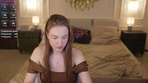 Video of a young woman with long brown hair, wearing a brown off-shoulder top, sitting on a bed with beige sheets. The background features a neatly made bed, two bedside lamps, and a floral wall art.