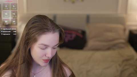 Video of a fair-skinned woman with long brown hair, wearing a pink top, sitting on a beige bed with pillows, in a softly lit bedroom.