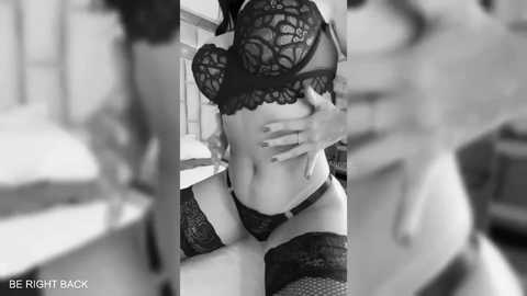Black-and-white video of a woman with light skin wearing black lace lingerie, including a bra and panties, and fishnet thigh-highs, standing indoors with a blurred background.