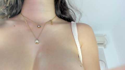 A video of a woman's upper body, showing her bare shoulders and a thin, white spaghetti strap top. She wears gold necklaces with small pendants. Her skin is light, and her dark hair falls over her shoulders.