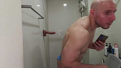 Video of a bald, muscular, white man in a shower, holding a smartphone, wearing a blue wristband. He is standing next to a wooden shower seat and a bottle rack.