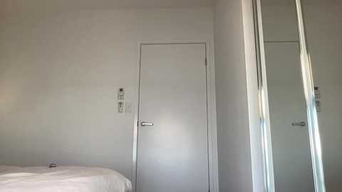 Video of a minimalist, modern bedroom with white walls, a door, and a large mirror reflecting the bed with white sheets.