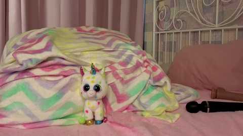 Video of a pink and white unicorn plushie under a rainbow-colored blanket, lying on a bed with a pink sheet and a white metal headboard. A black dildo lies on the bed beside the plushie.