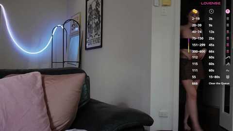 Video of a dimly lit room with a neon light, a black leather couch, and a person partially visible in a doorway, wearing black clothing.