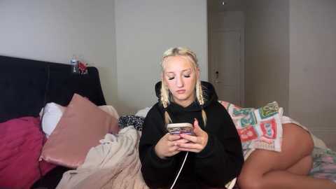 Video of a blonde woman in pajamas, sitting on a bed with messy blankets, engrossed in a smartphone, with a pink pillow and a colorful blanket in the background.
