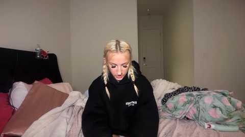Video of a blonde woman with braided hair in a messy bedroom, wearing a black hoodie, sitting on a bed with a pink and floral blanket.
