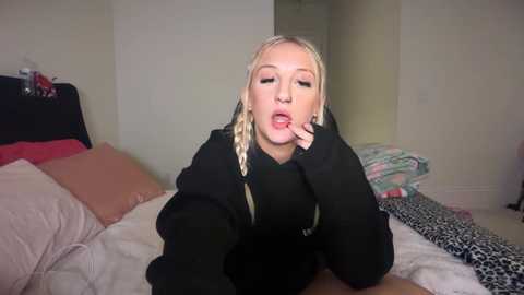 Video of a blonde woman with braided hair, wearing a black hoodie, lying on a bed with pink and white sheets, surrounded by pillows, and a leopard print blanket.