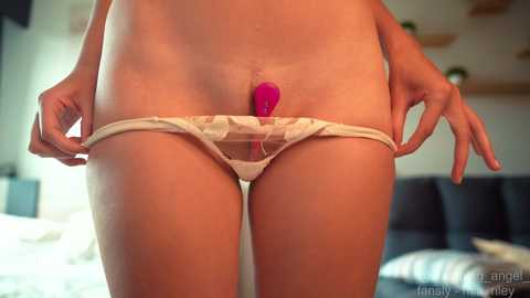 A video of a nude woman with a small pink dildo inserted into her vagina, wearing white lace panties. She is standing in a modern bedroom with a bed and wooden ceiling in the background.