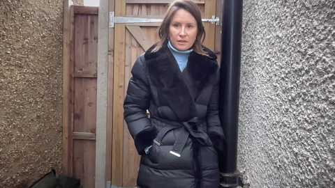 Video of a woman with shoulder-length brown hair, dressed in a black puffy coat with fur collar, standing in a narrow, rough-textured concrete alleyway next to a wooden door.