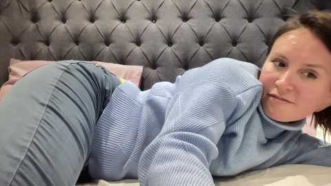 A video of a woman with light skin, dark hair, and a light blue knitted sweater lying on a bed with a tufted gray headboard and pink pillows.