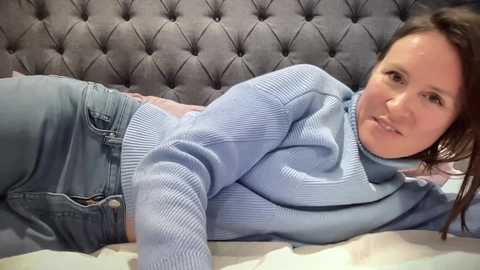 Video of a light-skinned woman with long brown hair, wearing a light blue knitted sweater and jeans, lying on a bed with a tufted gray headboard.