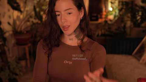 Video of a woman with long dark hair, wearing a brown \"Drip\" shirt, sitting indoors with potted plants and warm lighting, holding a drink.