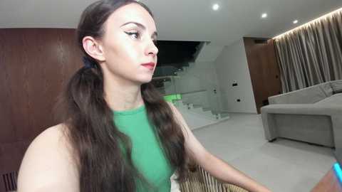Video of a young woman with long, dark hair in pigtails, wearing a green top, standing in a modern, minimalist living room with white walls, a grey couch, and wooden accents.